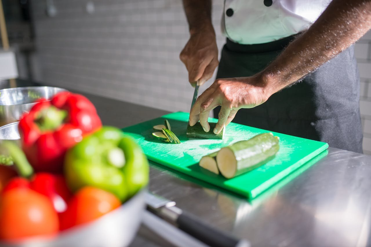 about-us Chef slicing zucchini on a green chopping board with fresh vegetables nearby.